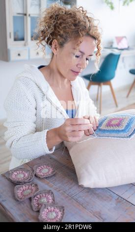 Happy adult woman sewing a pillow with handmade crochet squares sitting comfortably at the table at home. Indoor people leisure hobby activity concept lifestyle. Smiling lady working a home alone Stock Photo