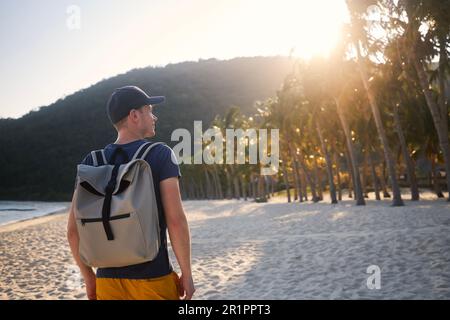 Rear view of tourist with backpack walking on beautiful sand beach with palm trees and looking at sunset. Stock Photo