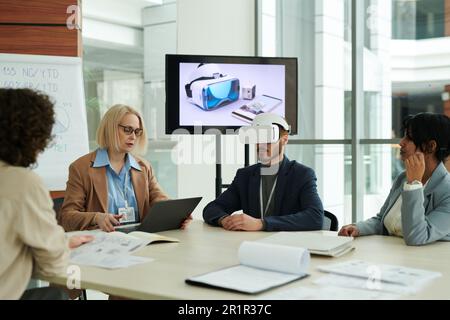 Young man with vr headset sitting by table among colleagues during presentation at meeting while mature woman using laptop Stock Photo