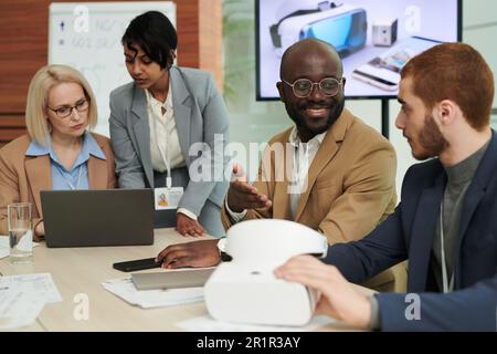 Happy young African American businessman talking to colleague with vr headset against two women discussing working points at meeting Stock Photo