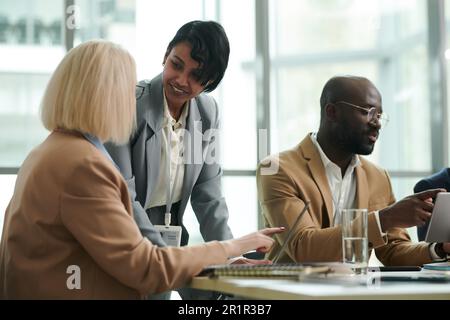 Young smiling businesswoman in formalwear looking at colleague pointing at laptop screen during discussion of online data at working meeting Stock Photo
