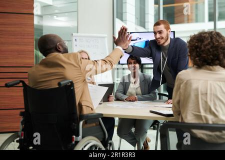 Two happy young intercultural businessmen giving each other high five over table while congratulating one another on successful project Stock Photo