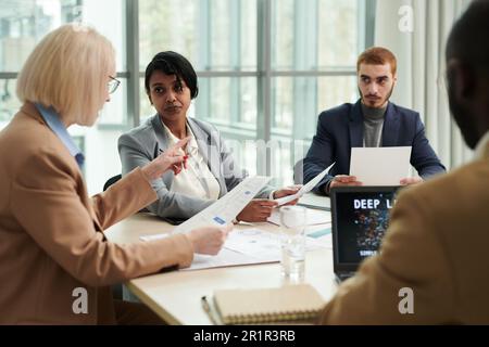 Blond female boss with financial document explaining something to group of young intercultural subordinates during discussion of data Stock Photo