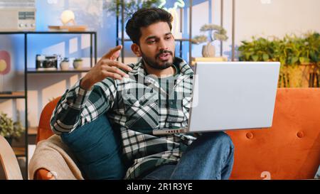Indian man sitting on couch looking at camera, making video webcam conference call with friends or family, enjoying pleasant conversation. Young hindu guy laughing waving hello at home room Stock Photo