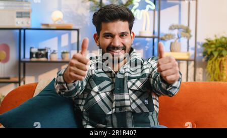 Like. Happy excited indian man looking approvingly at camera showing thumbs up, like sign positive something, good great news, positive feedback. Young hindu guy sitting on couch at home living room Stock Photo