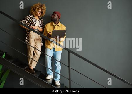 Two young economists or analysts discussing online graphic data on staircase while one of them making presentation of financial charts Stock Photo
