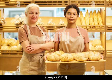 Two intercultural female bakers or clerks of bakery shop in uniform looking at camera while standing by display with assortment of fresh bread Stock Photo
