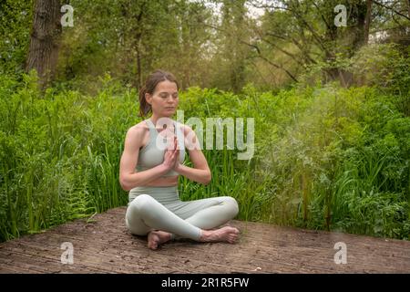 middle aged fit healthy woman practicing yoga outside in natural calm green environment in park meditating with folded palms in front her Stock Photo