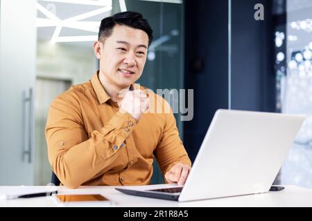 Cheerful and smiling Asian man working inside office, businessman in shirt and glasses using laptop, engineer developer programmer typing code programming software Stock Photo