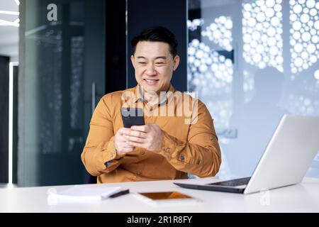 Young handsome Asian man, office worker sitting in the office at the table, fiddling with the phone, typing messages, chatting Stock Photo