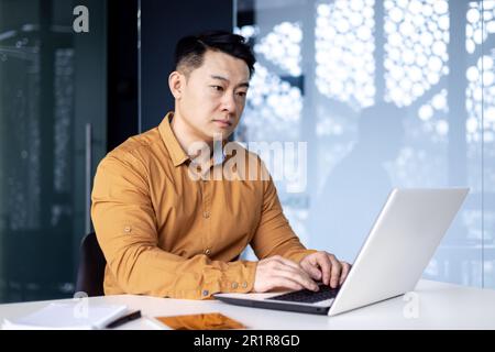 Serious and thoughtful young man of Asian origin programmer, developer works in office at desktop at laptop. Stock Photo