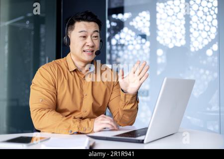 Successful smiling man working inside office using laptop and headset for video call, asian cheerfully consulting tech support customer, businessman at work in shirt. Stock Photo