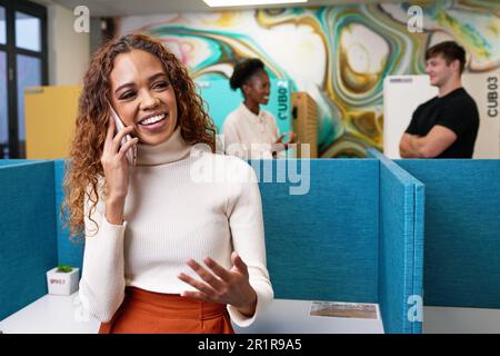 Young multi-ethnic woman talks on phone in open plan office with colleagues Stock Photo