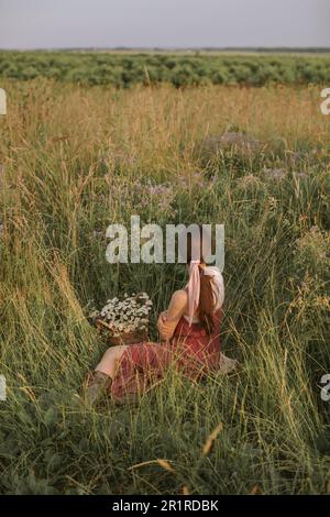 Rear view of a woman sitting in a meadow in spring with a basket of daisies, Belarus Stock Photo