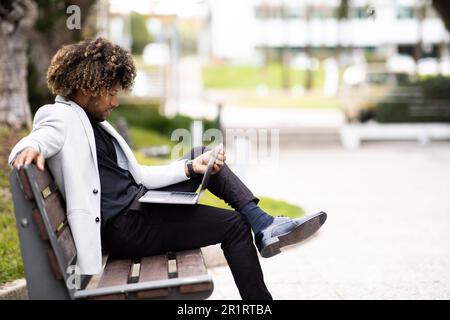 Black businessman in suit using laptop while sitting on bench near modern office building, side view, free space Stock Photo