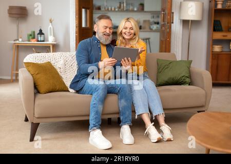 Cheerful Elderly Spouses Using Tablet Sitting On Couch At Home Stock Photo
