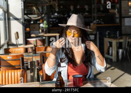 Stylish woman in sunglasses and hat relaxing sitting in a coffee shop. Stock Photo