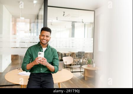 Positive handsome indian male office employee enjoying spending time online in social networks, standing in office and using smartphone, freelancer man holds mobile phone messaging and chatting Stock Photo