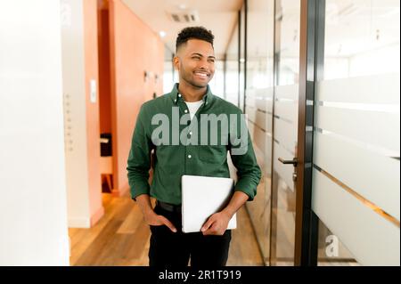 Waist up portrait of the proud indian businessman carrying laptop in hand and smiling, Latin freelancer man posing in modern loft office with glass partition on the background, looks aside Stock Photo