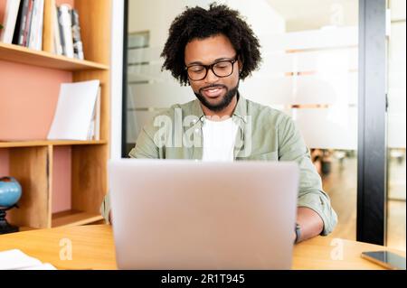 African-American guy using laptop for remote work sitting in the modern coworking space, bookshelves on the background, man in casual wear and glasses typing indoor Stock Photo