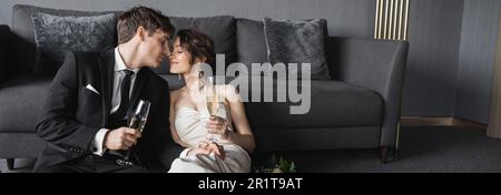 bride in white wedding dress and groom in black suit holding glasses of champagne while kissing and celebrating their marriage near bridal bouquet aft Stock Photo