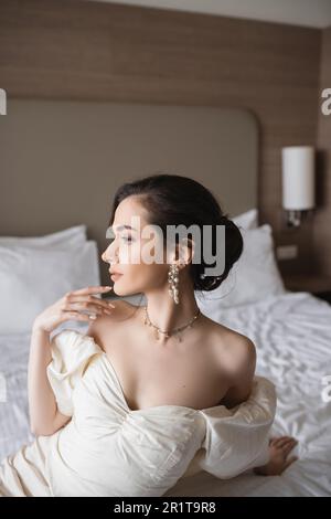 side view of enchanting young bride in white dress and luxurious jewelry with pearl earrings and necklace looking away in modern bedroom in hotel room Stock Photo