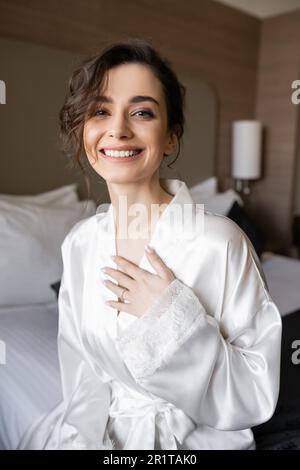 happy young bride with brunette hair and engagement ring on finger smiling while sitting in white silk robe and looking at camera in hotel room on wed Stock Photo