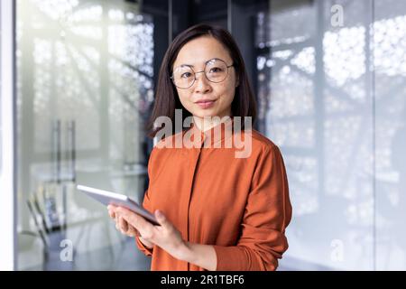 Portrait of a young Asian woman programmer, developer, tester in glasses standing in the office, holding a tablet, working online and looking, posing at the camera. Stock Photo