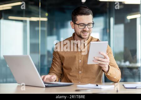A young man works in an office center, sits at a table with a laptop, holds a tablet in his hand and talks on a video call with a smile. Stock Photo