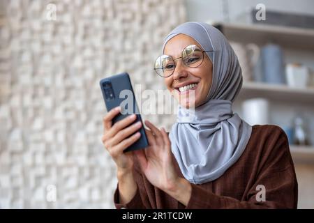Close-up photo. Young Muslim student woman in black hijab sitting at home and using mobile phone smiling. Stock Photo
