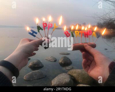 Burning happy birthday inscription made of holiday candles in the hands of a man and a woman opposite the water of the ocean lake river. Concept: birt Stock Photo