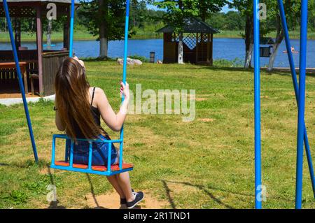 Beautiful young woman girl with long hair rides a swing in the park on the nature on the shore of the lake in summer. Stock Photo