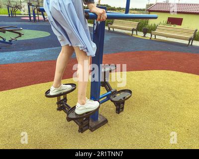 doing sports on the simulator in the park in the fresh air. a girl in a blue dress and white sneakers trains her legs on the platform. active leisure, Stock Photo