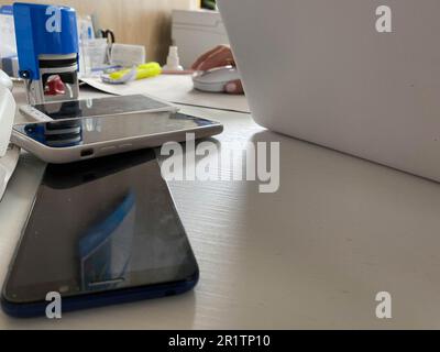 Two working touchscreen mobile phones, smartphones lie on the table in the office with stationery, a stapler, a seal and a laptop. Stock Photo