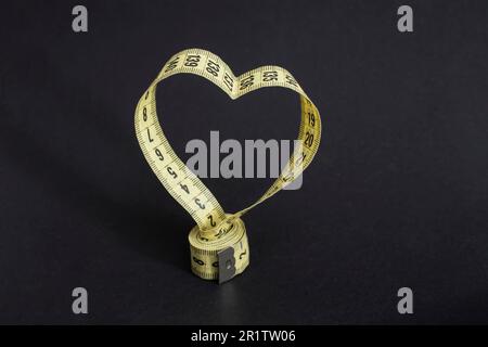 Rolled yellow tape measure in the shape of a heart, isolated on black background. Stock Photo