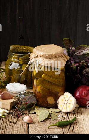 Pickled cucumbers in closed and open jars, onion, garlic, dill, parsley, basil on a dark wooden background. Homemade canned gherkins , spices and herb Stock Photo