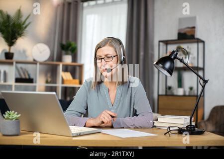 Middle-aged elegant woman using headset with microphone while enjoying home comfort during remote work. Smiling caucasian freelancer in eyeglasses holding online videoconference and making notes. Stock Photo