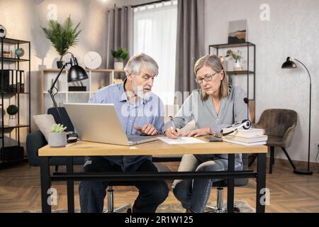 Middle-aged lady in glasses putting down information while senior male finding data on laptop in creative office. Efficient professionals drafting written agreement while doing distant work from home. Stock Photo