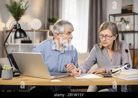 Middle-aged lady in glasses putting down information while senior male finding data on laptop in creative office. Efficient professionals drafting written agreement while doing distant work from home. Stock Photo
