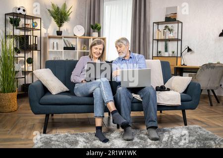Serious mature people in casual clothes using laptops while spending time in cozy environment at home. Elderly family man and wife reading online article on digital screen while sitting on couch. Stock Photo