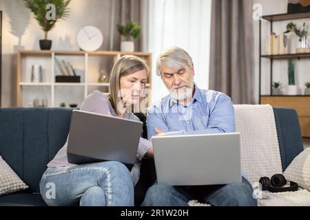 Serious mature people in casual clothes using laptops while spending time in cozy environment at home. Elderly family man and wife reading online article on digital screen while sitting on couch. Stock Photo