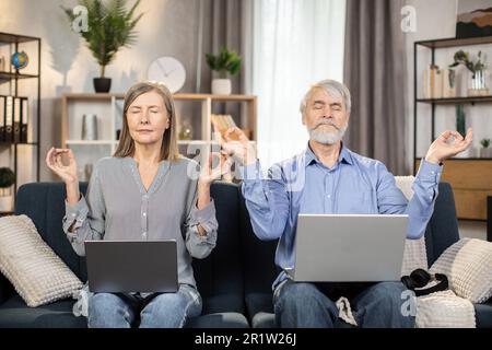 Portrait of aged spouses holding fingers in gyan mudra while relaxing on couch with laptops on knees in room interior. Gray-haired man and woman reducing fatigue using meditation practice at home. Stock Photo