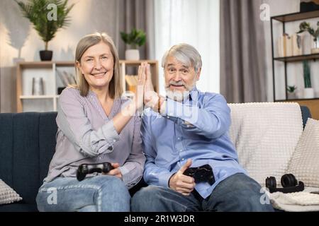 Friendly caucasian adults of mature age giving gentle high five after relaxing with video games via controllers at home. Happy spouses getting great pleasure of spending time together during weekend. Stock Photo