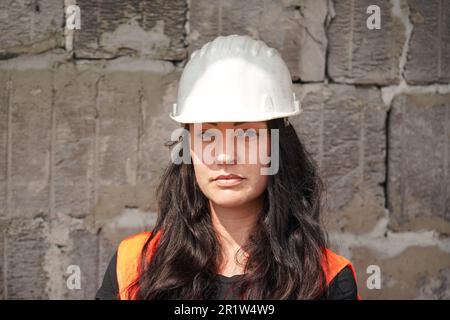 Young woman in white hard hat and orange high visibility vest, long dark hair, looking into camera. Blurred old construction site wall background Stock Photo