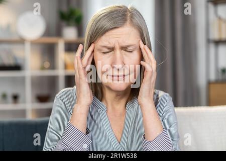Frustrated senior lady suffering from strong sudden migraine and dizziness while sitting on couch. Sick aged woman in striped shirt massaging temples with fingers indoors. Concept of healthcare. Stock Photo