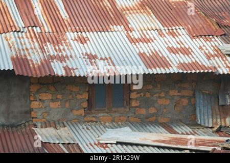 close-up view of old damaged village shanty house, temporary building made of unbaked brick wall with cement and rusty metal corrugated roofing sheets Stock Photo