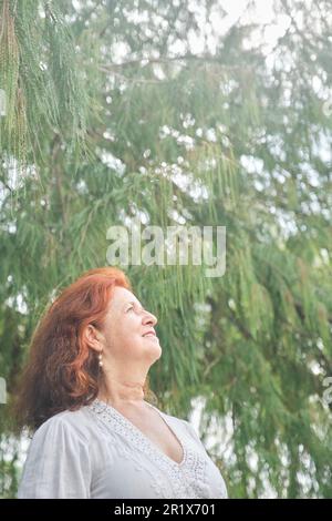 Portrait of a latin senior red-haired woman smiling looking up daydreaming in nature. Bright composition with copy space. Concept: peace of mind and h Stock Photo