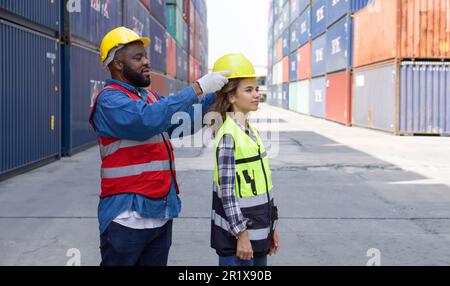 A foreman with mustache and beard helps a female apprentice wear a hardhat. Both of them wearing reflective safety vest. There are container in the wo Stock Photo