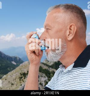 Mature man using asthma inhaler in mountains. Emergency first aid during outdoor recreation Stock Photo