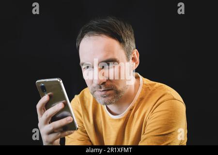 Portrait of cheerful brunette man in black shirt using cellphone and smiling, reading good news message, enjoying mobile application. Stock Photo